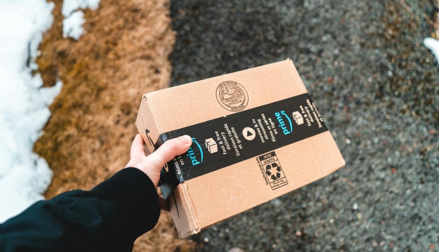 A person holding a cardboard Amazon Prime package on a snowy urban sidewalk.